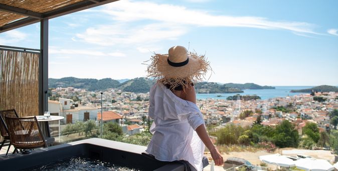Eine Frau mit Sonnenhut sitzt auf dem Rand eines Whirlpools auf einem Balkon und blickt runter Richtung der Stadt