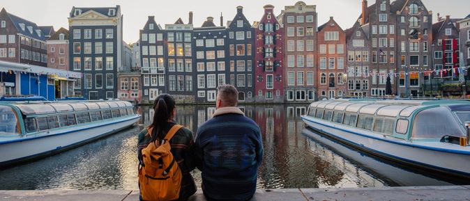 Zwei Personen sitzen in Amsterdam am Wasser und schauen gemeinsam auf die gegenüberliegenden bunten Häuser und die Boote