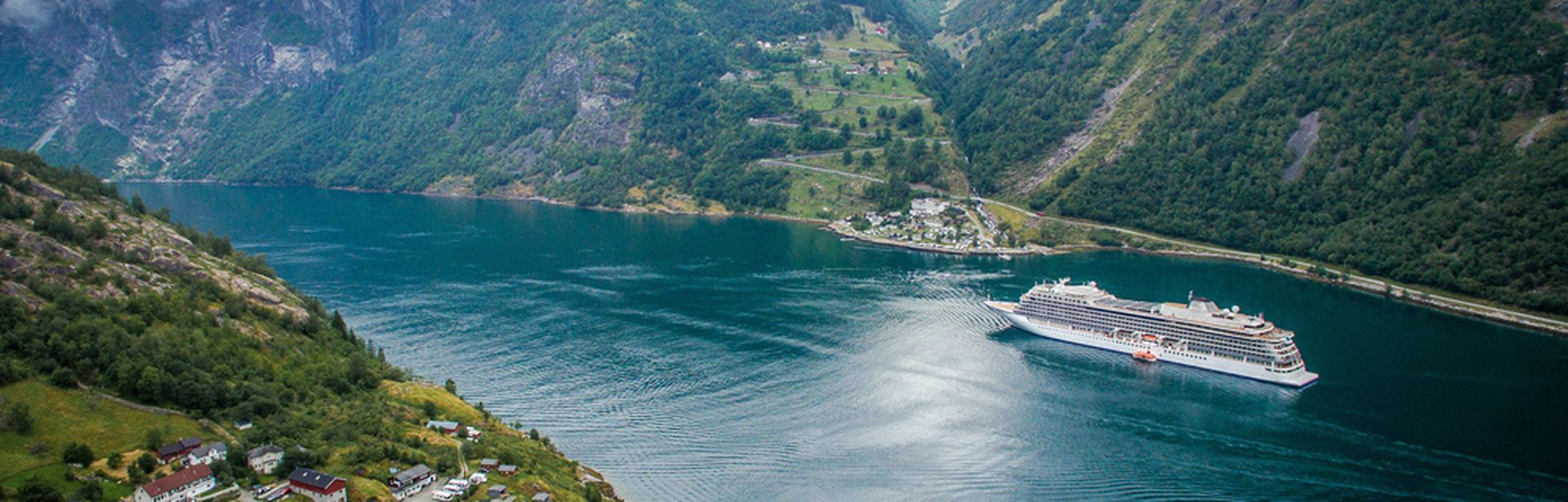 Ein Kreuzfahrtschiff fährt durch einen tiefen, von grünen Bergen gesäumten Fjord mit einem kleinen Dorf im Vordergrund.