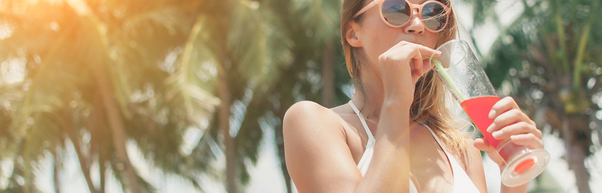 Eine Frau im weißen Bikini und einer Sonnenbrille trinkt mit einem Strohhalm einen roten Cocktail, im Hintergrund sind Palmen zu sehen