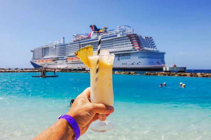 Eine Hand hält einen Cocktail hoch, im Hintergrund ist ein großes Kreuzfahrtschiff