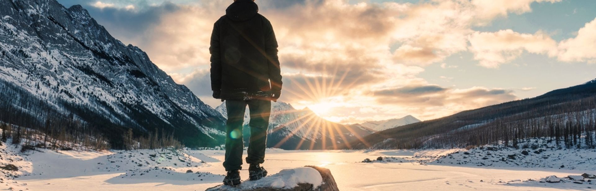 Mann steht leicht erhöht auf einem großen Stein in den Bergen bei Schnee und schaut in die untergehende Sonne