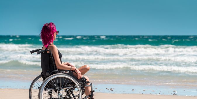 Frau im Rollstuhl mit einem Bein sitzt entspannt am Strand und blickt aufs Meer