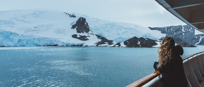 Eine Frau steht mit wehenden Haaren an der Reling eines Kreuzfahrtschiffes und schaut auf schneebedeckte Berge