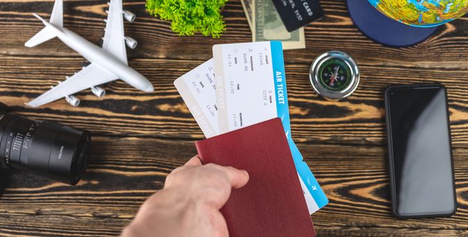 Auf einem Holztisch liegen ein Handy, ein Miniaturflugzeug, eine Kamera, ein Kompass, Geld und ein Globus. Eine Hand hält einen Reisepass und zwei Flugtickets