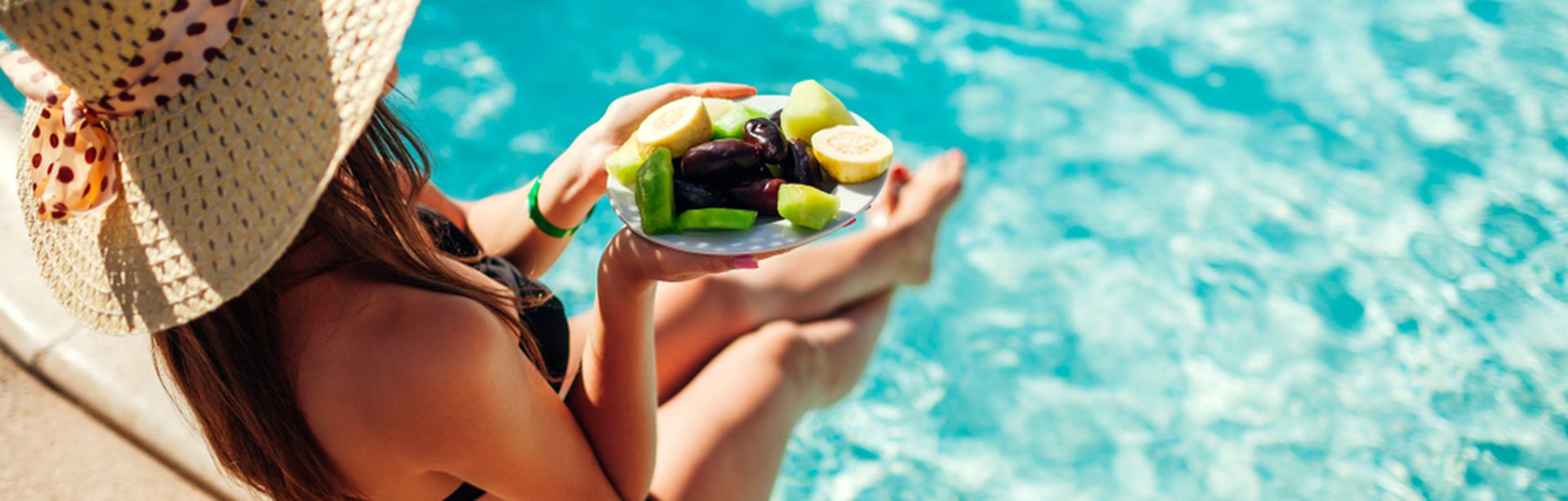Eine Frau von oben fotografiert, sie trägt einen Strohhut und sitzt im Bikini am Pool, in der Hand hat sie einen Teller mit geschnittenem Obst