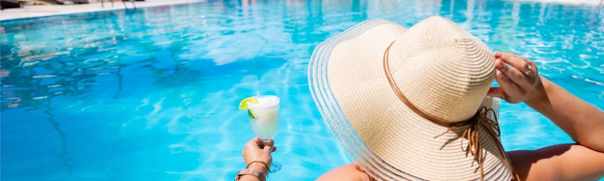 Eine Frau mit Hut von hinten, sie sitzt in einem Pool und hat ein weißes Getränk in der Hand