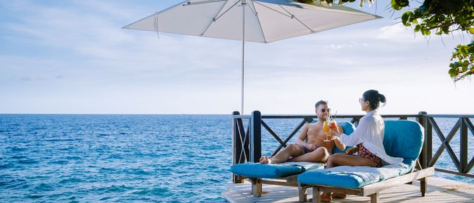 Eine Frau und ein Mann sitzen unter einem Sonnenschirm auf Liegen auf einem Steg und stoßen mit Cocktails an