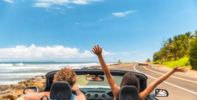 Zwei Personen fahren in einem Auto eine Straße am Meer entlang, eine von ihnen reißt die Arme nach oben