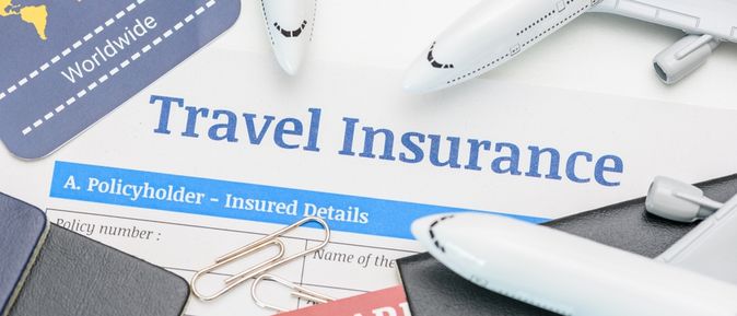 In der Mitte des Bildes steht "Travel Insurance" darum liegen mehrere Büroklammern, Reisepässe und mehrere kleine Flugzeuge