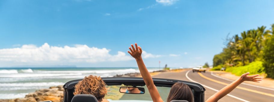 Zwei Personen fahren in einem Auto eine Straße am Meer entlang, eine von ihnen reißt die Arme nach oben