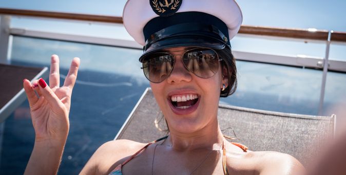 Eine Frau im Bikini und einer Kapitänsmütze auf macht ein Selfie und zeigt lächelnd ein Peace Zeichen in die Kamera
