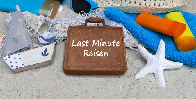 Im Sand liegen ein kleines flaches Holzboot, ein weißer Seestern, ein Sonnenhut, ein Handtuch und ein brauner Koffer mit der Aufschrift "Last Minute Reisen"