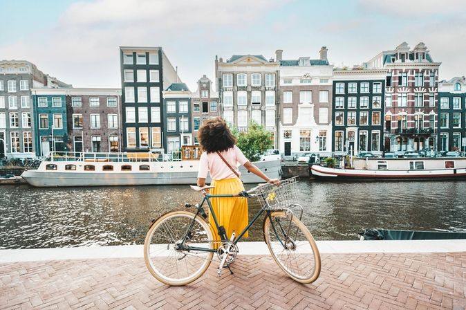 Eine Frau in einem gelben Rock steht an ihrem Fahrrad und schaut auf die Amstel und die gegenüberliegenden Häuser und Boote in Amsterdam