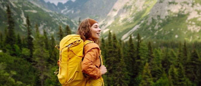 Eine Frau mit gelbem Wanderrucksack steht auf einem Berg und schaut begeistert in die Ferne