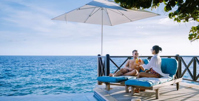 Eine Frau und ein Mann sitzen unter einem Sonnenschirm auf Liegen auf einem Steg und stoßen mit Cocktails an