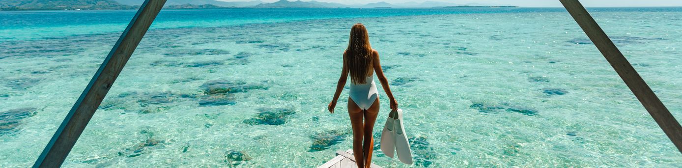 Eine Frau im Bikini steht am Ende eines Stegs und schaut aufs Meer