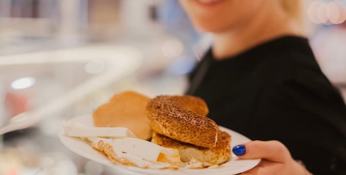 Eine lächelnde Frau hält einen Teller mit Brötchen, Käse und anderem Frühstück in die Kamera