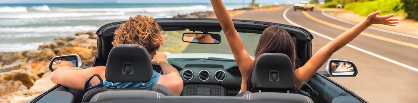 Zwei Personen im Auto mit offenem Verdeck, im Hintergrund kann man das Meer sehen