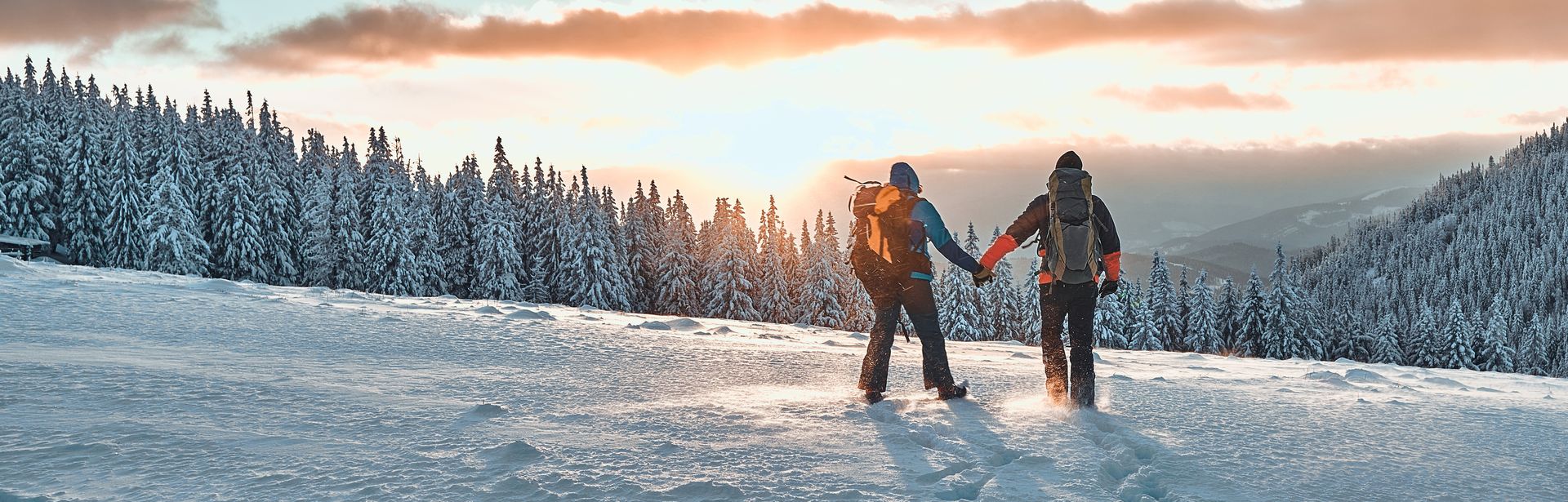 Zwei Personen stehen auf einem Schneebedeckten Berg und schauen Händchen haltend in den Sonnenuntergang