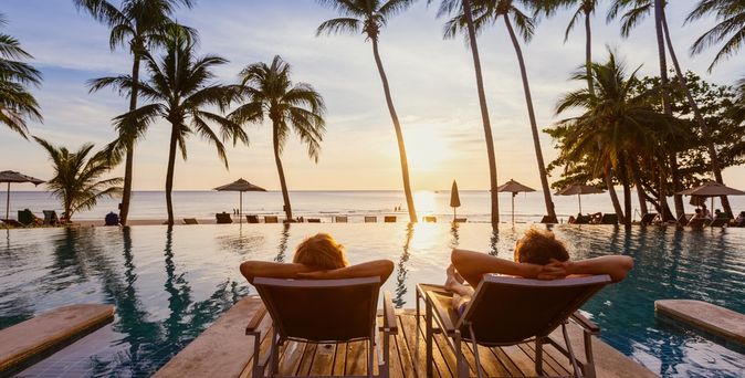 Zwei Personen von hinten fotografiert, Sie liegen auf zwei Liegen auf einem Steg im Pool und schauen auf den Sonnenuntergang über dem Meer