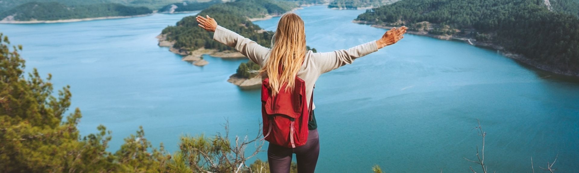 Eine Frau mit Rucksack schaut von einer Klippe auf einen großen See, eine Berglandschaft und breitet die Arme aus