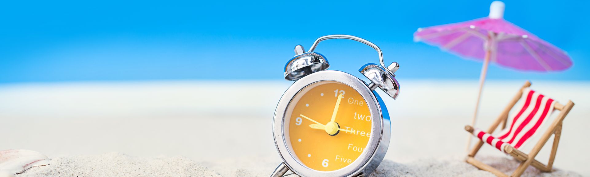 Im Sand stehen ein gelber Wecker, eine Miniatur Liege mit Sonnenschirm und einige Muscheln