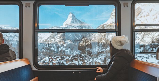 Eine Frau mit Winterjacke und Mütze sitzt in einem Zug und schaut aus dem Fenster in die Schneebedeckte Landschaft