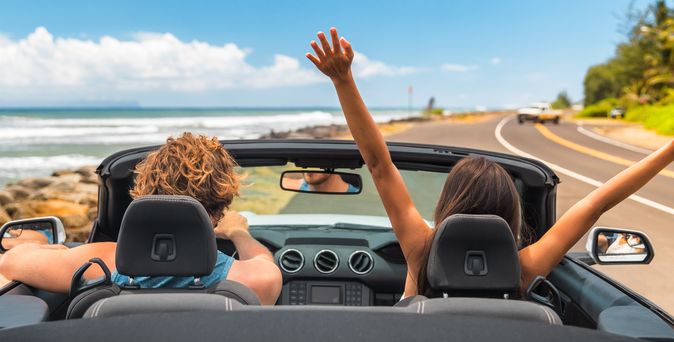 Zwei Personen im Auto mit offenem Verdeck, im Hintergrund kann man das Meer sehen