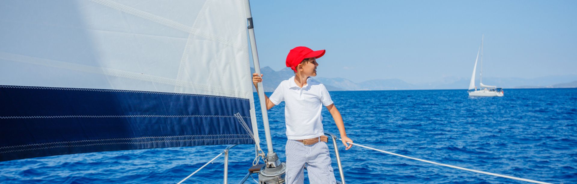 Junge mit roter Kappe steht vorne auf einem Segelboot und schaut auf das Meer