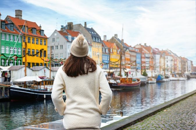 Eine Frau von hinten. Sie trägt eine weiße Jacke und eine Mütze. Sie schaut auf bunte Häuser, das Wasser und einige Boote in Nyhavn in Kopenhagen.