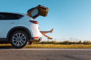 Ein Auto ist zur Hälfte von der Seite zu sehen und steht am Straßenrand an einer Wiese. Der Kofferraum ist auf und man sieht Beine herausragen