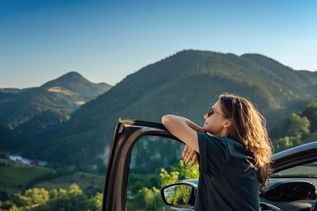 Eine Frau lehnt sich lächelnd auf die offene Tür eines Autos und schaut in die Natur um sie herum