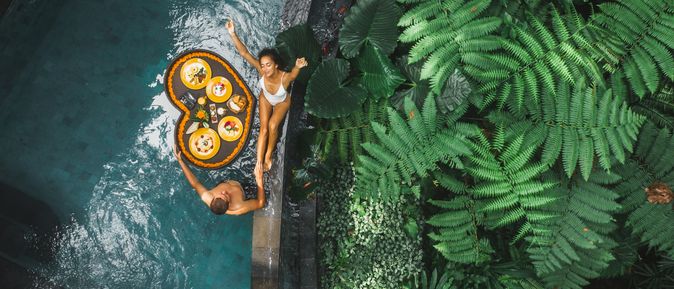 Ein Paar von oben, eine Frau sitzt am Rand eines Pools, ein Mann steht im Pool, zwischen ihnen schwimmt ein Herzförmiges Tablett mit Essen