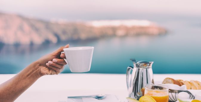 Ein gedeckter Tisch mit einer Kanne und Tellern mit Obst, eine Hand hält eine Tasse hoch, im Hintergrund ist das Meer