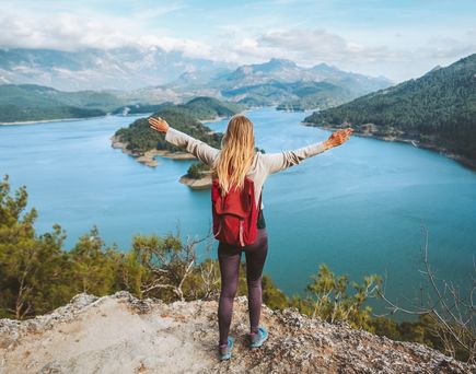 Eine Frau mit Rucksack schaut von einer Klippe auf einen großen See, eine Berglandschaft und breitet die Arme aus