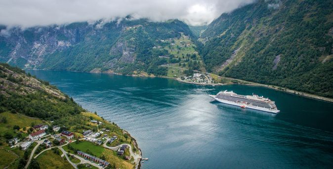 Ein Kreuzfahrtschiff fährt durch einen tiefen, von grünen Bergen gesäumten Fjord mit einem kleinen Dorf im Vordergrund.