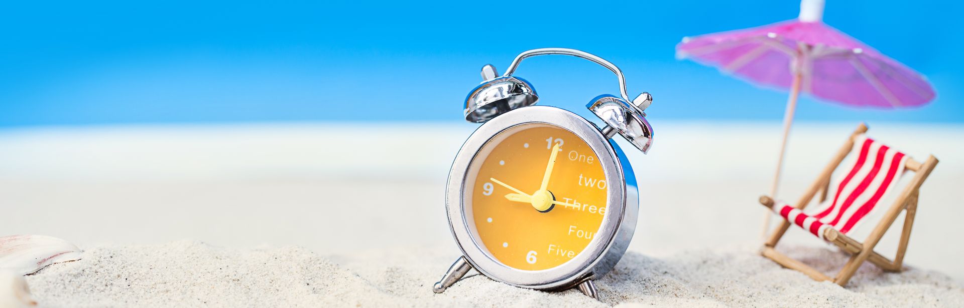 Im Sand stehen ein gelber Wecker, eine Miniatur Liege mit Sonnenschirm und einige Muscheln