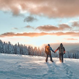 Zwei Personen stehen auf einem Schneebedeckten Berg und schauen Händchen haltend in den Sonnenuntergang
