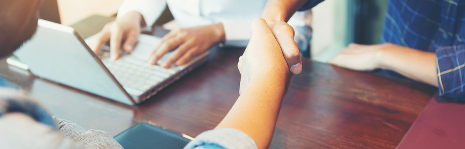 Zwei Personen geben sich über einem Schreibtisch die Hand und daneben sitzt eine weitere Person mit Laptop an dem Schreibtisch
