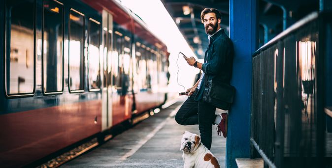 Ein Mann und ein Hund stehen gemeinsam an einem Bahnsteig und schauen in die Kamera