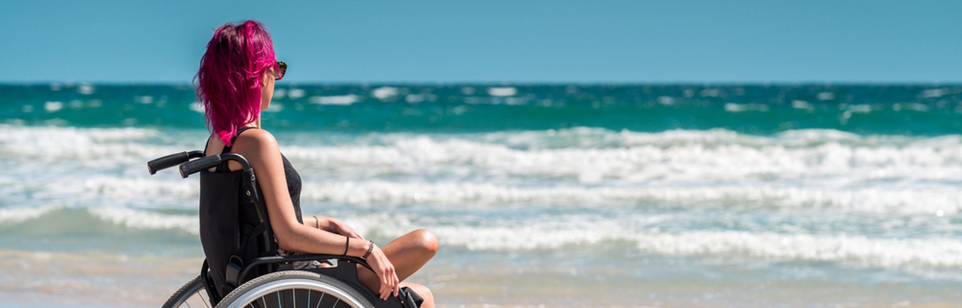 Frau im Rollstuhl mit einem Bein sitzt entspannt am Strand und blickt aufs Meer