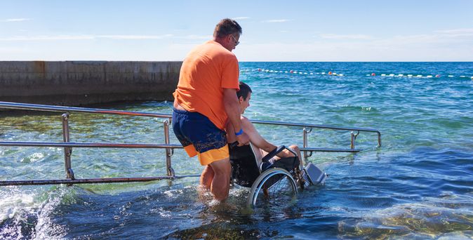 Mann hilft einem anderen Mann im Rollstuhl über eine Treppe ins Meer