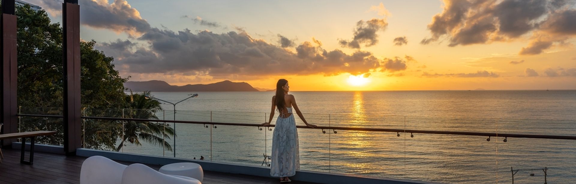 Frau am Geländer einer Terasse schaut auf den Sonnenuntergang über dem Meer