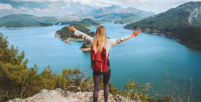 Eine Frau trägt einen roten Rucksack, streckt die Arme aus und schaut von einem Berg runter auf einen See mit Insel