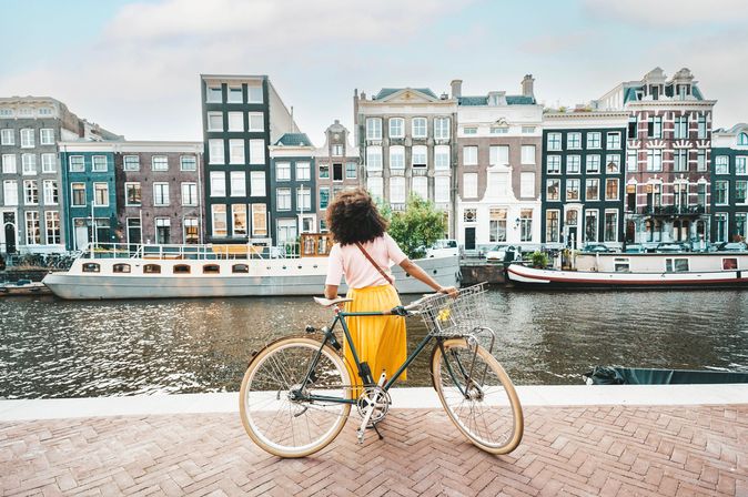 Eine Frau steht mit ihrem Fahrrad vor der Amstel und schaut auf die Häuser auf der gegenüberliegenden Seite