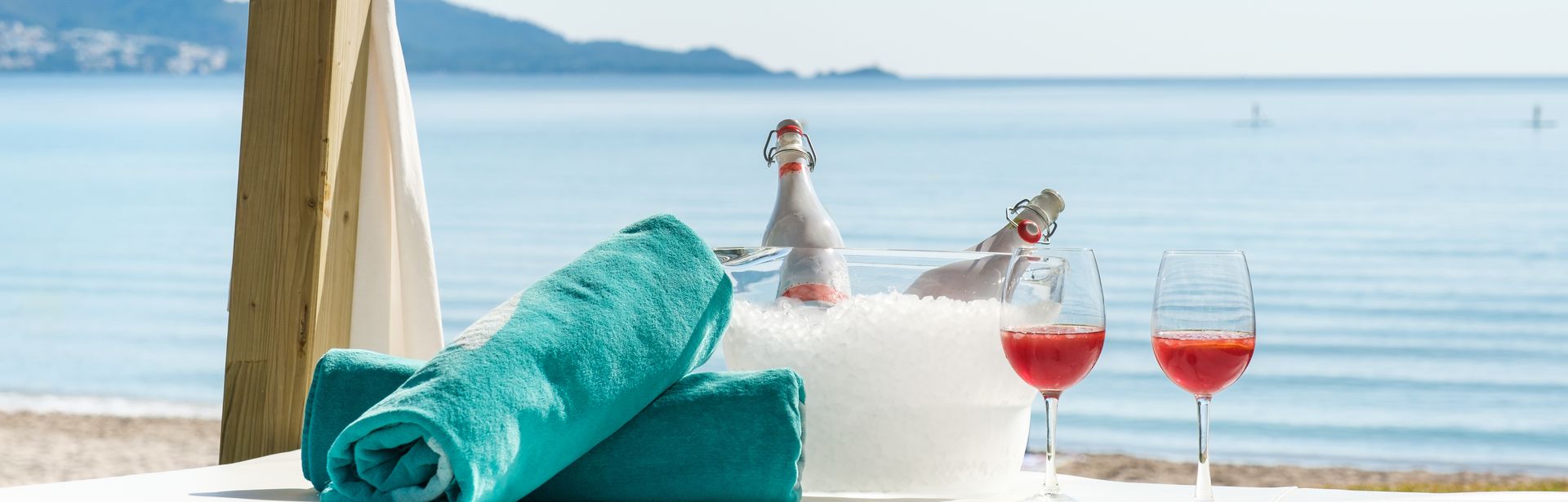 Zwei aufgerollte Handtücher liegen neben zwei Gläsern und ein Getränkekühler mit Eis und zwei Flaschen auf einer Decke am Strand