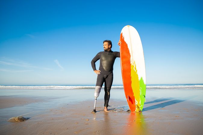 Ein Mann mit einer Beinprothese am linken Bein steht in einem Neoprenanzug am Strand und hält ein buntes Surfboard