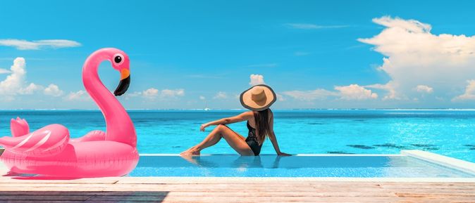 Frau sitzt am Rand eines Pools mit Meerblick, neben Ihr ein aufblasbarer Flamingo