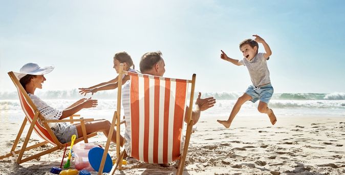 Zwei Personen sitzen in Liegestühlen am Strand, zwei Kinder laufen lachend auf sie zu